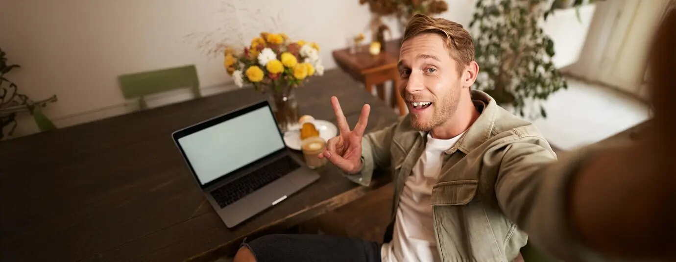 Selfie-Porträt eines gutaussehenden jungen Mannes, der in einem Café mit Laptop ein Foto von sich selbst macht und lächelt.