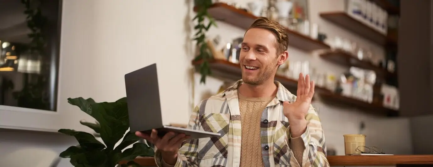 Bild eines jungen Mannes im Café, der sich per Laptop in einen Videoanruf einwählt, online mit Freunden spricht und der Kamera zur Begrüßung zuwinkt.